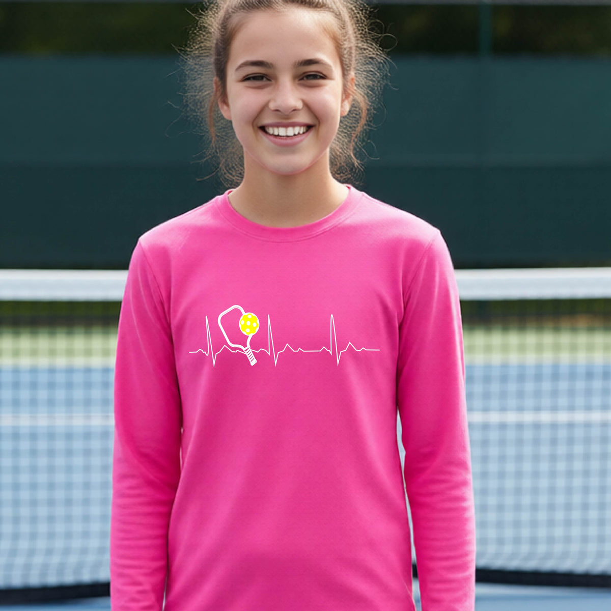 A close-up of a young person wearing bright pink long-sleeve t-shirt showing a white pickleball graphic of a heartbeat EKG including a pickleball paddle and a yellow pickleball, in front of a pickleball court.