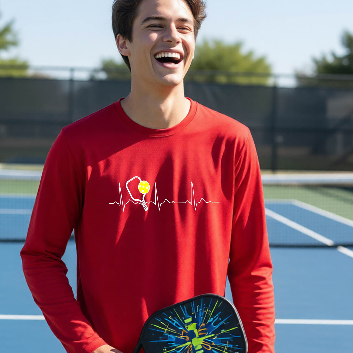 A close-up of a young person wearing a red long-sleeve t-shirt showing a white pickleball graphic of a heartbeat EKG including a pickleball paddle and a yellow pickleball, in front of a pickleball court.