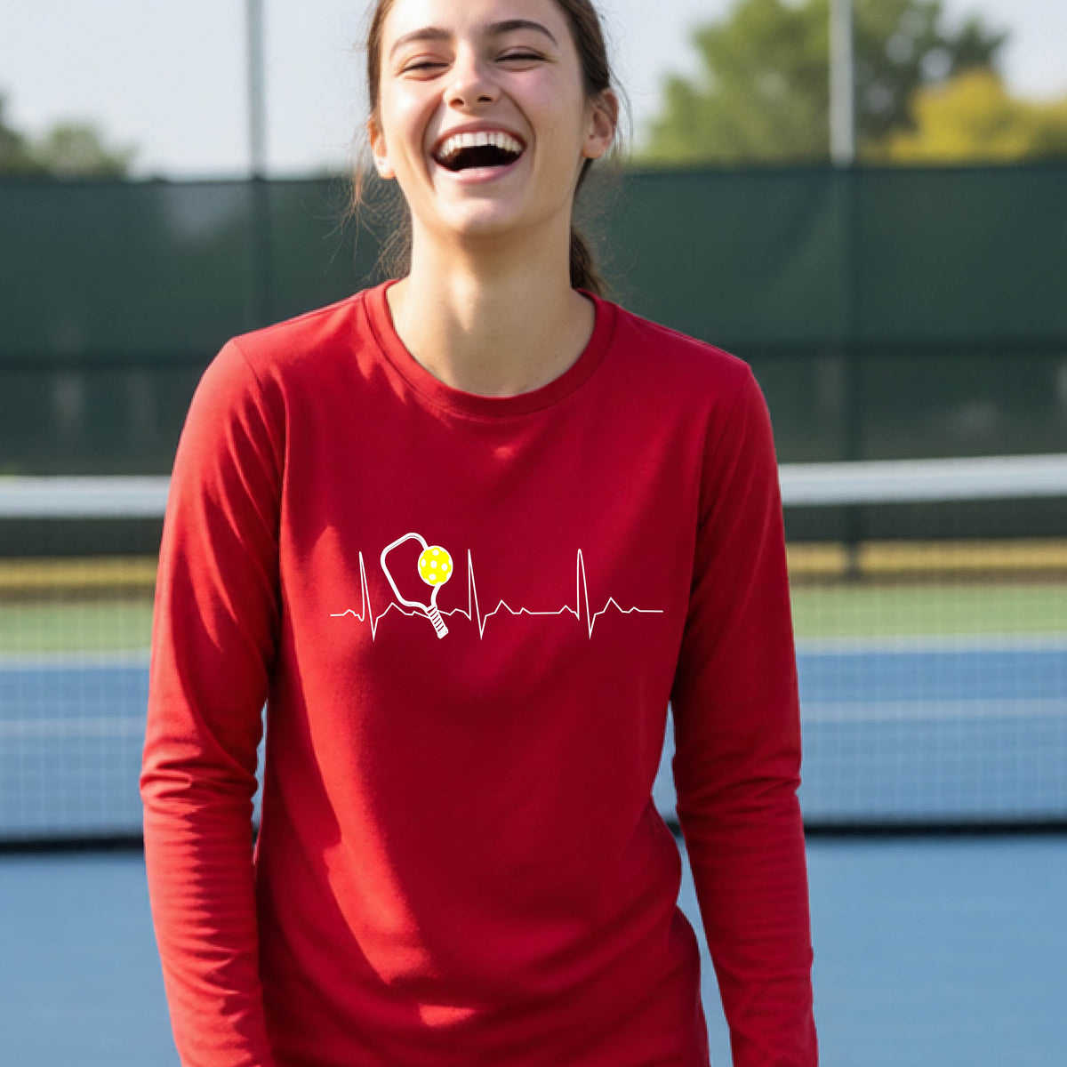 A close-up of a young person wearing red long-sleeve t-shirt showing a white pickleball graphic of a heartbeat EKG including a pickleball paddle and a yellow pickleball, in front of a pickleball court.
