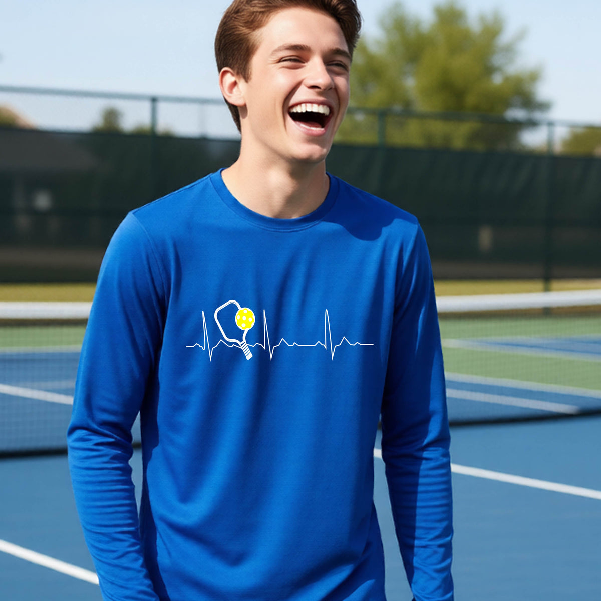 A close-up of a young person wearing royal blue long-sleeve t-shirt showing a white pickleball graphic of a heartbeat EKG including a pickleball paddle and a yellow pickleball, in front of a pickleball court.