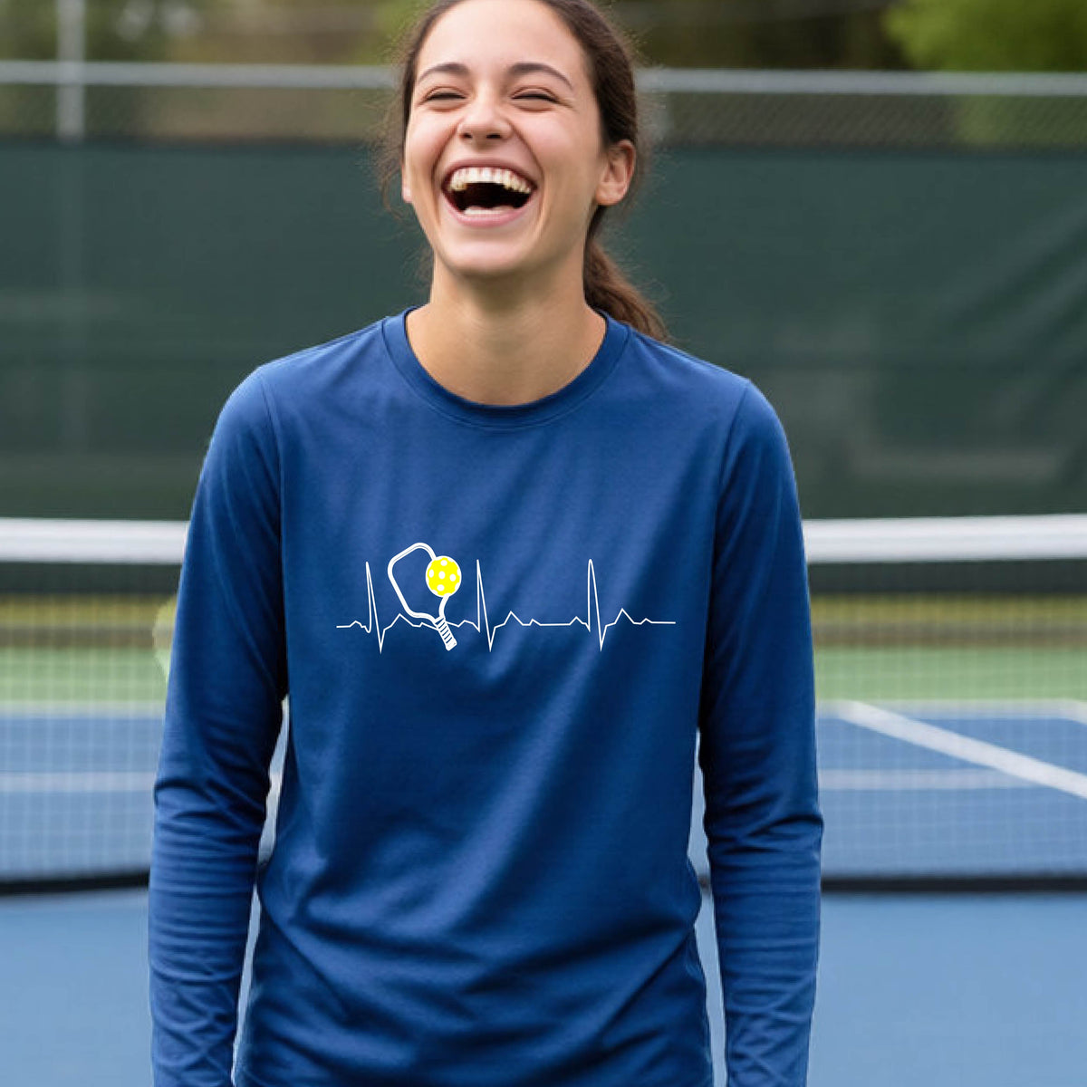 A close-up of a young person wearing royal long-sleeve t-shirt showing a white pickleball graphic of a heartbeat EKG including a pickleball paddle and a yellow pickleball, in front of a pickleball court.