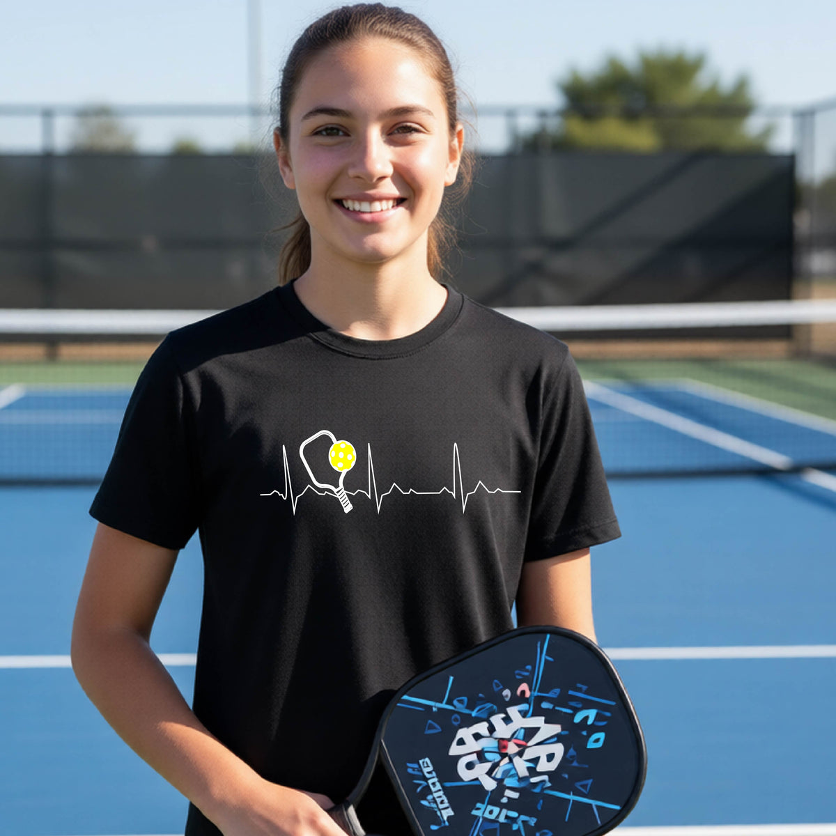 A close-up of a young person in a black short-sleeve t-shirt sporting a white pickleball graphic of a heartbeat EKG including a pickleball paddle and a yellow pickleball across the check, in front of a pickleball court.