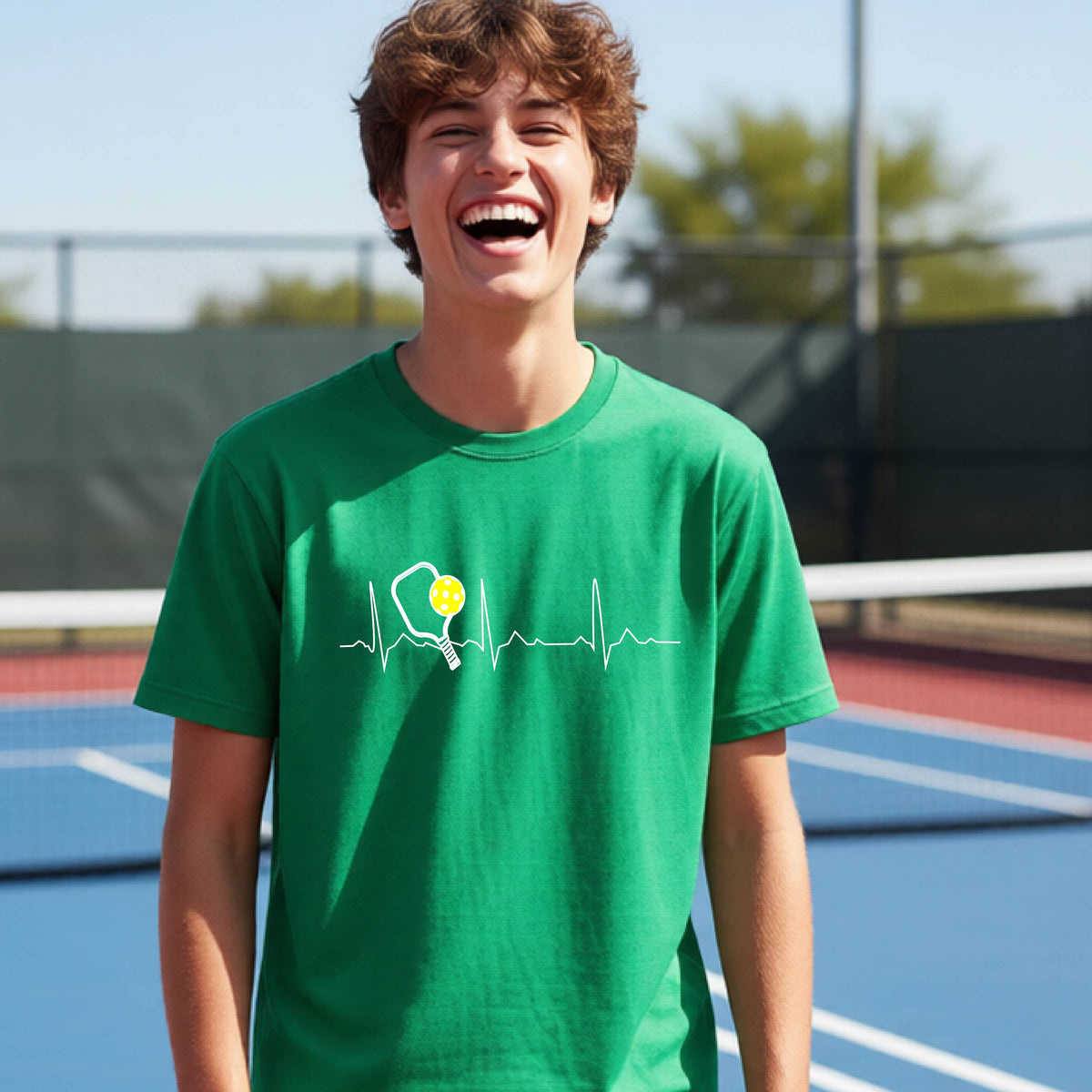 A close-up of a young person in a green short-sleeve t-shirt sporting a white pickleball graphic of a heartbeat EKG including a pickleball paddle and a yellow pickleball across the check, in front of a pickleball court.