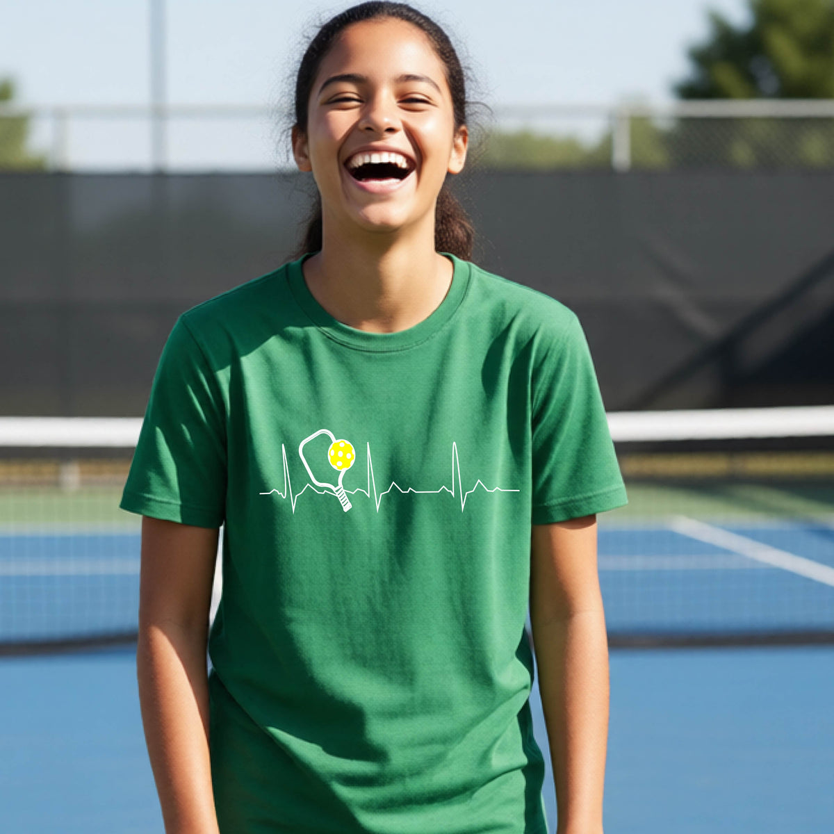 A close-up of a young person in a green short-sleeve t-shirt sporting a white pickleball graphic of a heartbeat EKG including a pickleball paddle and a yellow pickleball across the check, in front of a pickleball court.