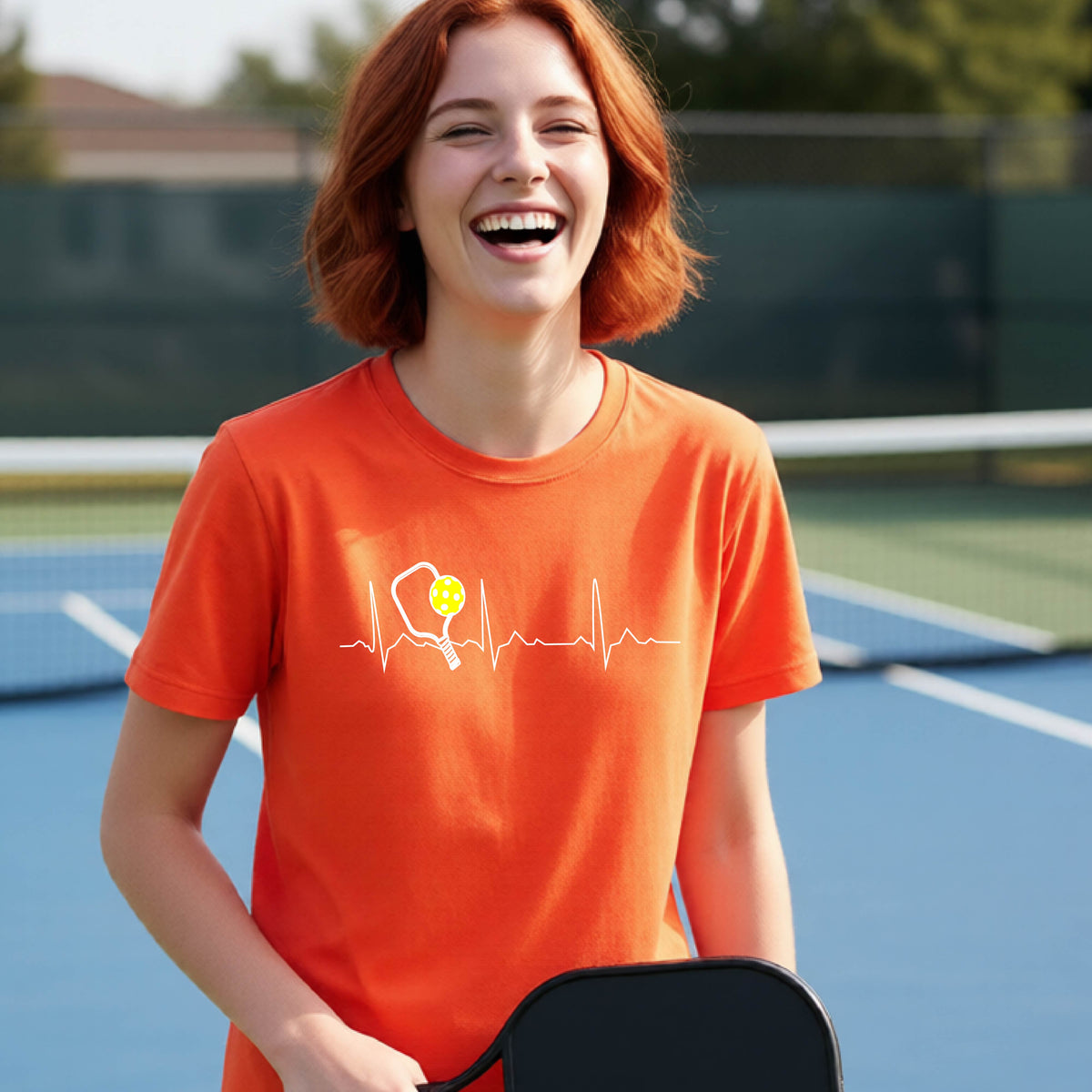 A close-up of a young person in an orange short-sleeve t-shirt sporting a white pickleball graphic of a heartbeat EKG including a pickleball paddle and a yellow pickleball across the check, in front of a pickleball court.