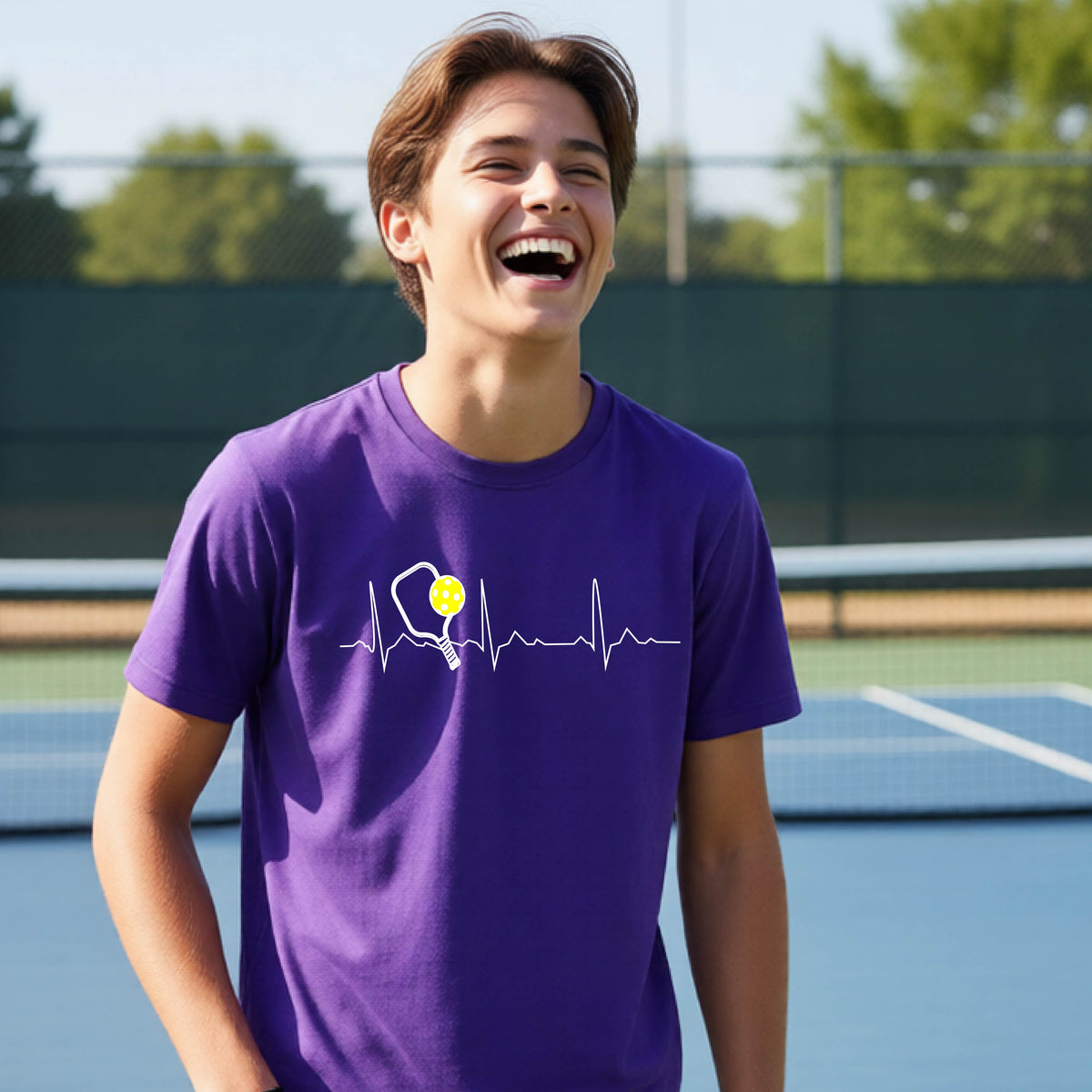 A close-up of a young person in a purple short-sleeve t-shirt sporting a white pickleball graphic of a heartbeat EKG including a pickleball paddle and a yellow pickleball across the check, in front of a pickleball court.