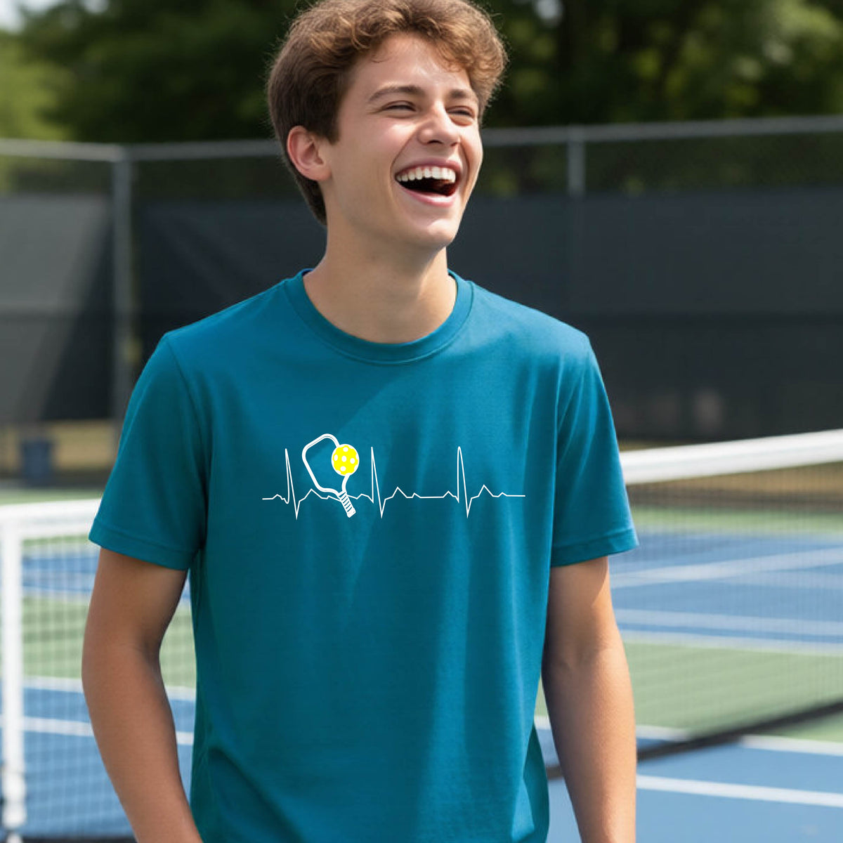 A close-up of a young person in a teal short-sleeve t-shirt sporting a white pickleball graphic of a heartbeat EKG including a pickleball paddle and a yellow pickleball across the check, in front of a pickleball court.