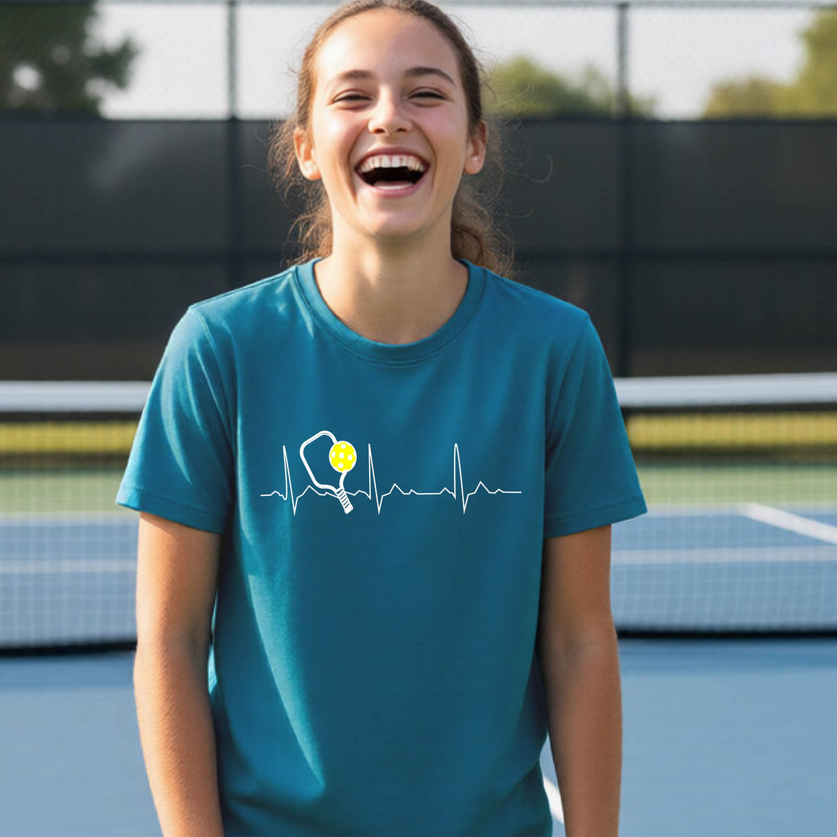 A close-up of a young person in a teal short-sleeve t-shirt sporting a white pickleball graphic of a heartbeat EKG including a pickleball paddle and a yellow pickleball across the check, in front of a pickleball court.