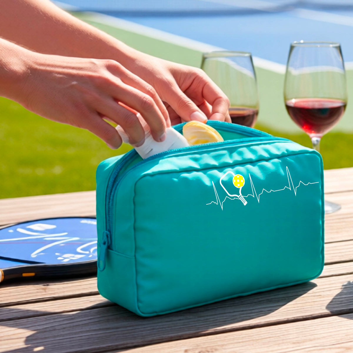 A close-up of a turquoise blue pickleball cosmetic toiletry bag showing a white pickleball graphic of a heartbeat EKG including a pickleball paddle and a yellow pickleball, in front of a pickleball court.