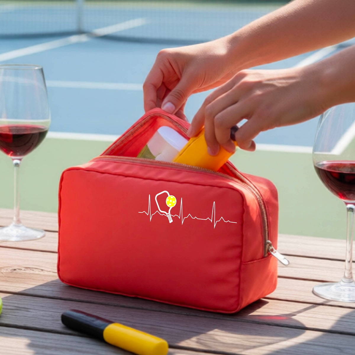 A close-up of a coral pickleball cosmetic toiletry bag showing a white pickleball graphic of a heartbeat EKG including a pickleball paddle and a yellow pickleball, in front of a pickleball court.