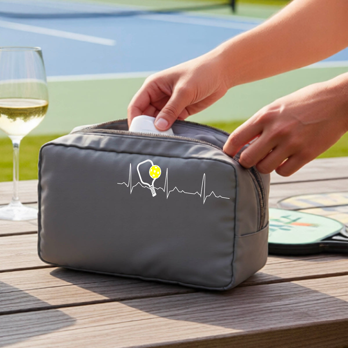 A close-up of a gray pickleball cosmetic toiletry bag showing a white pickleball graphic of a heartbeat EKG including a pickleball paddle and a yellow pickleball, in front of a pickleball court.