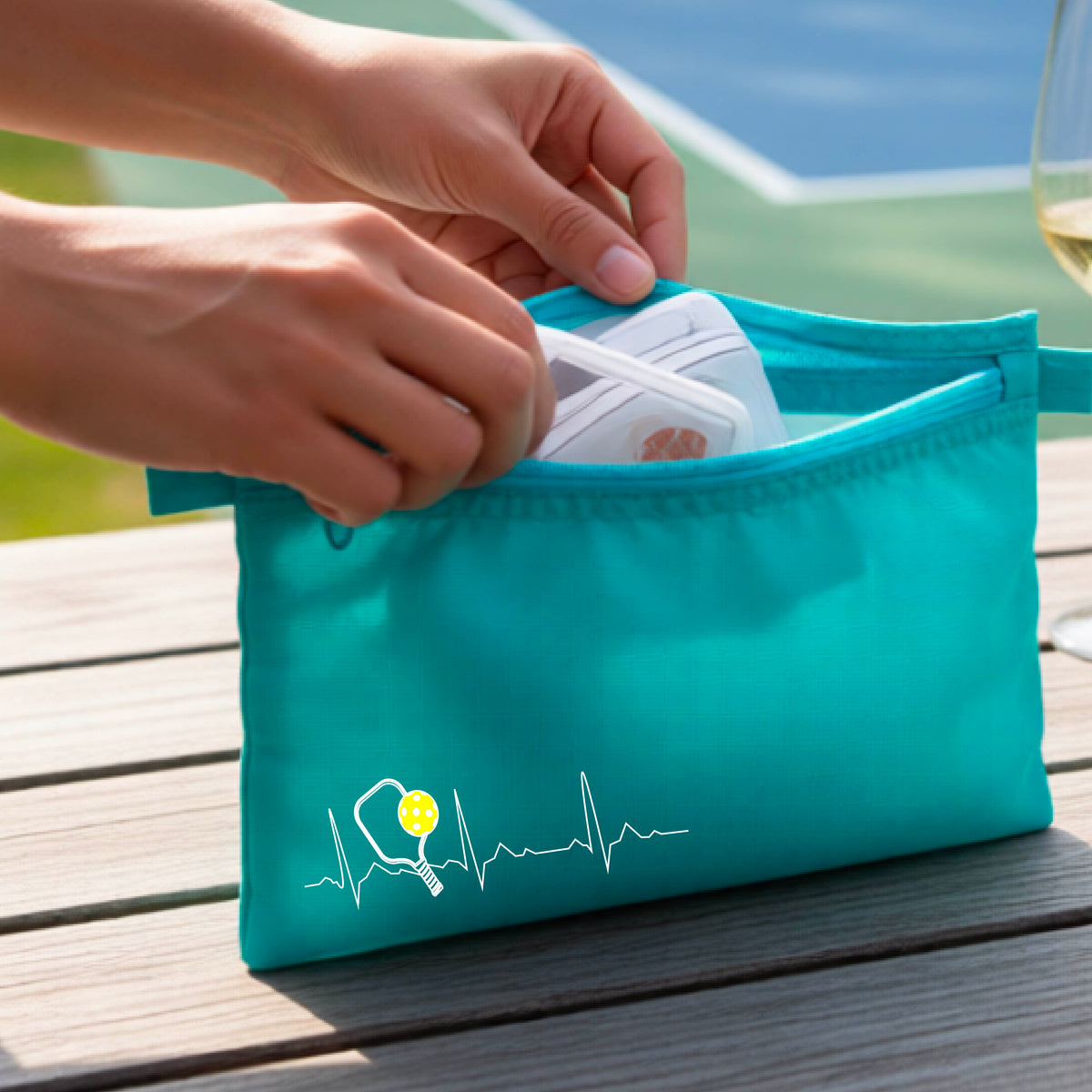 A close-up of a turquoise cosmetic toiletry bag showing a white pickleball graphic of a heartbeat EKG including a pickleball paddle and a yellow pickleball, in front of a pickleball court.