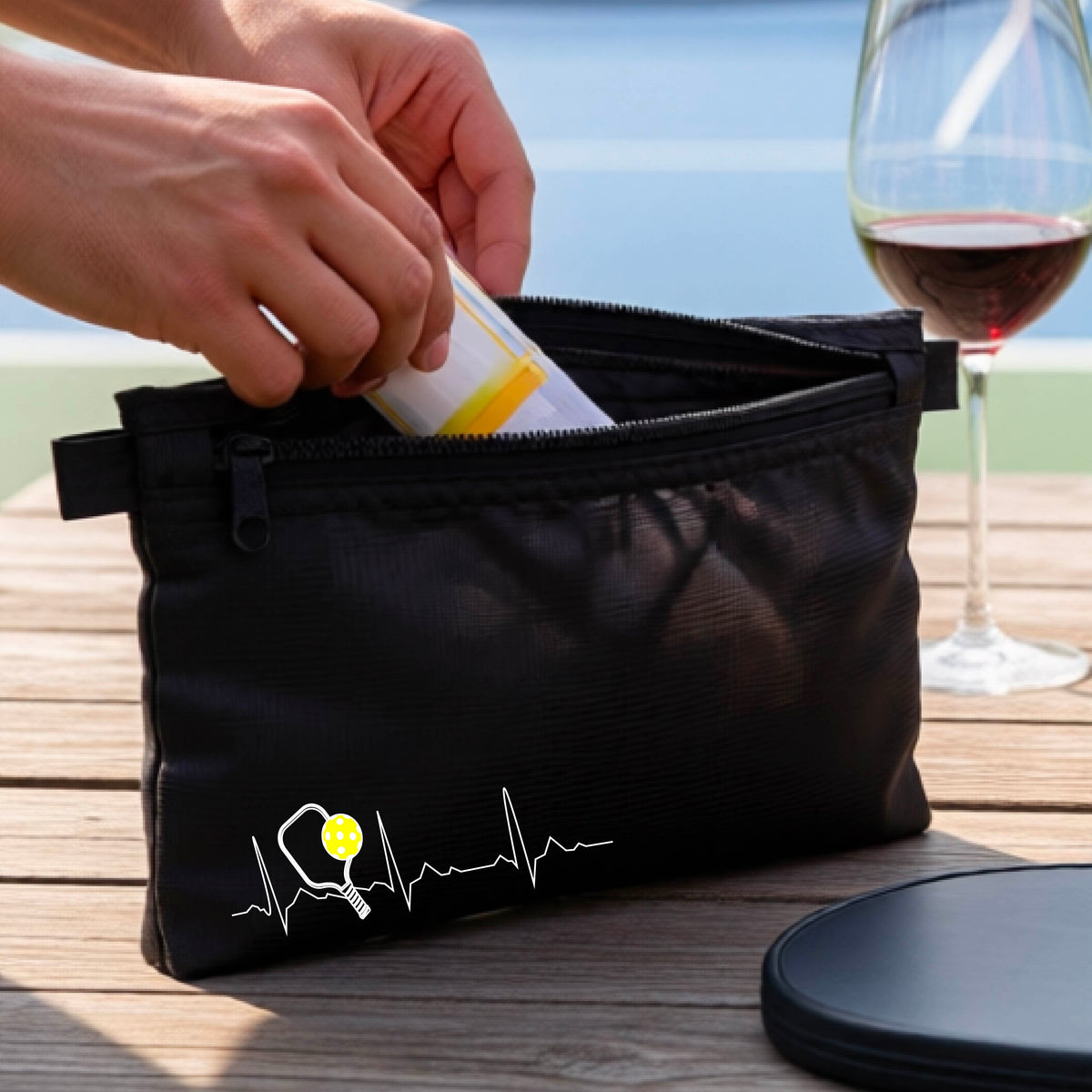 A close-up of a black cosmetic toiletry bag showing a white pickleball graphic of a heartbeat EKG including a pickleball paddle and a yellow pickleball, in front of a pickleball court.