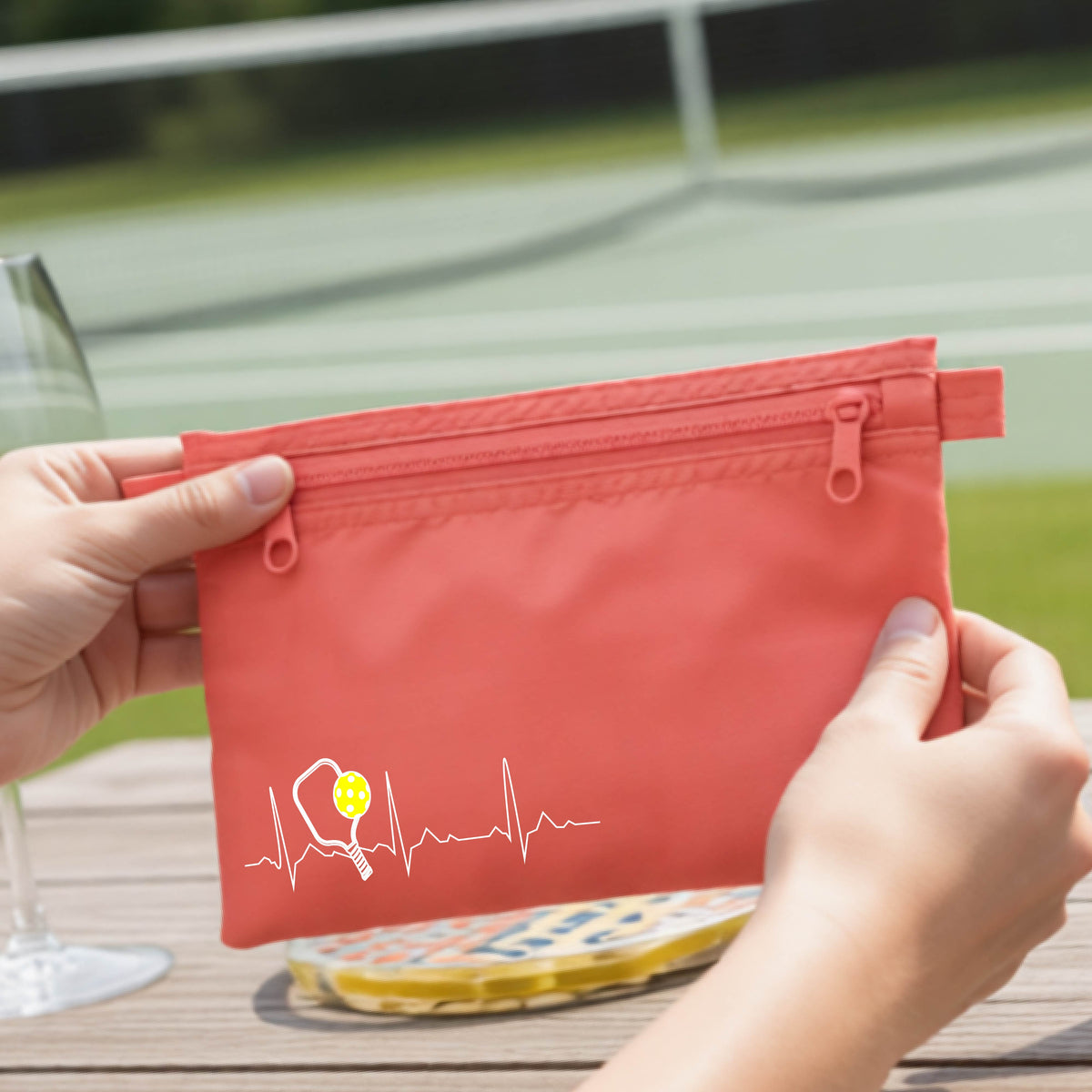 A close-up of a coral cosmetic toiletry bag showing a white pickleball graphic of a heartbeat EKG including a pickleball paddle and a yellow pickleball, in front of a pickleball court.