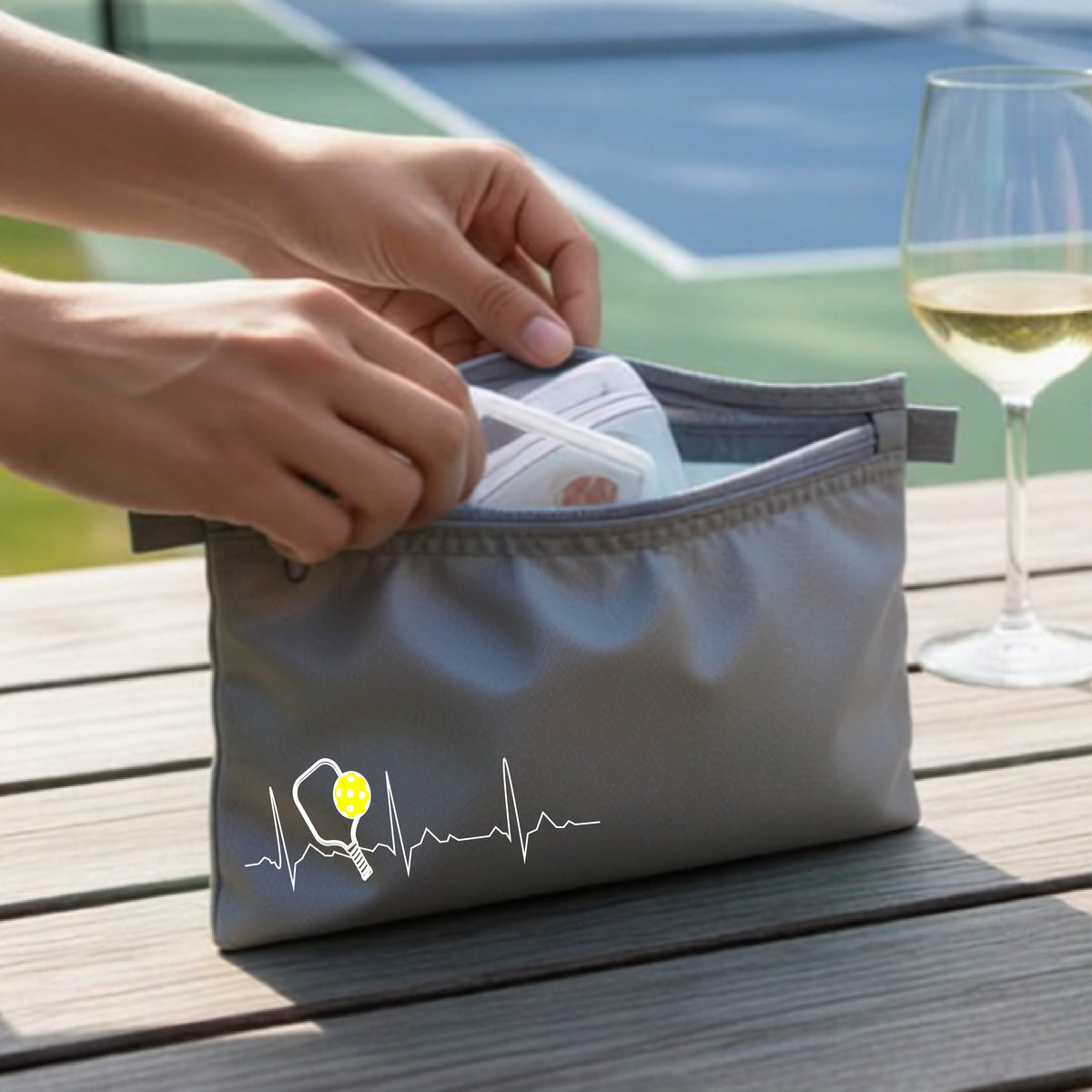 A close-up of a gray cosmetic toiletry bag showing a white pickleball graphic of a heartbeat EKG including a pickleball paddle and a yellow pickleball, in front of a pickleball court.