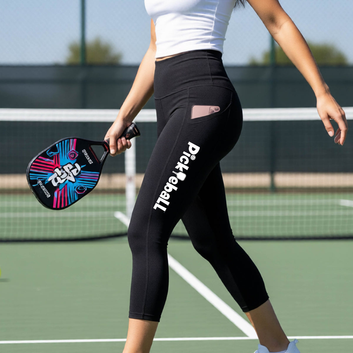 A close-up lifestyle shot of a woman on a pickleball court wearing high-waisted black capris with 'Pickleball' printed in white down the side of her her leg. 