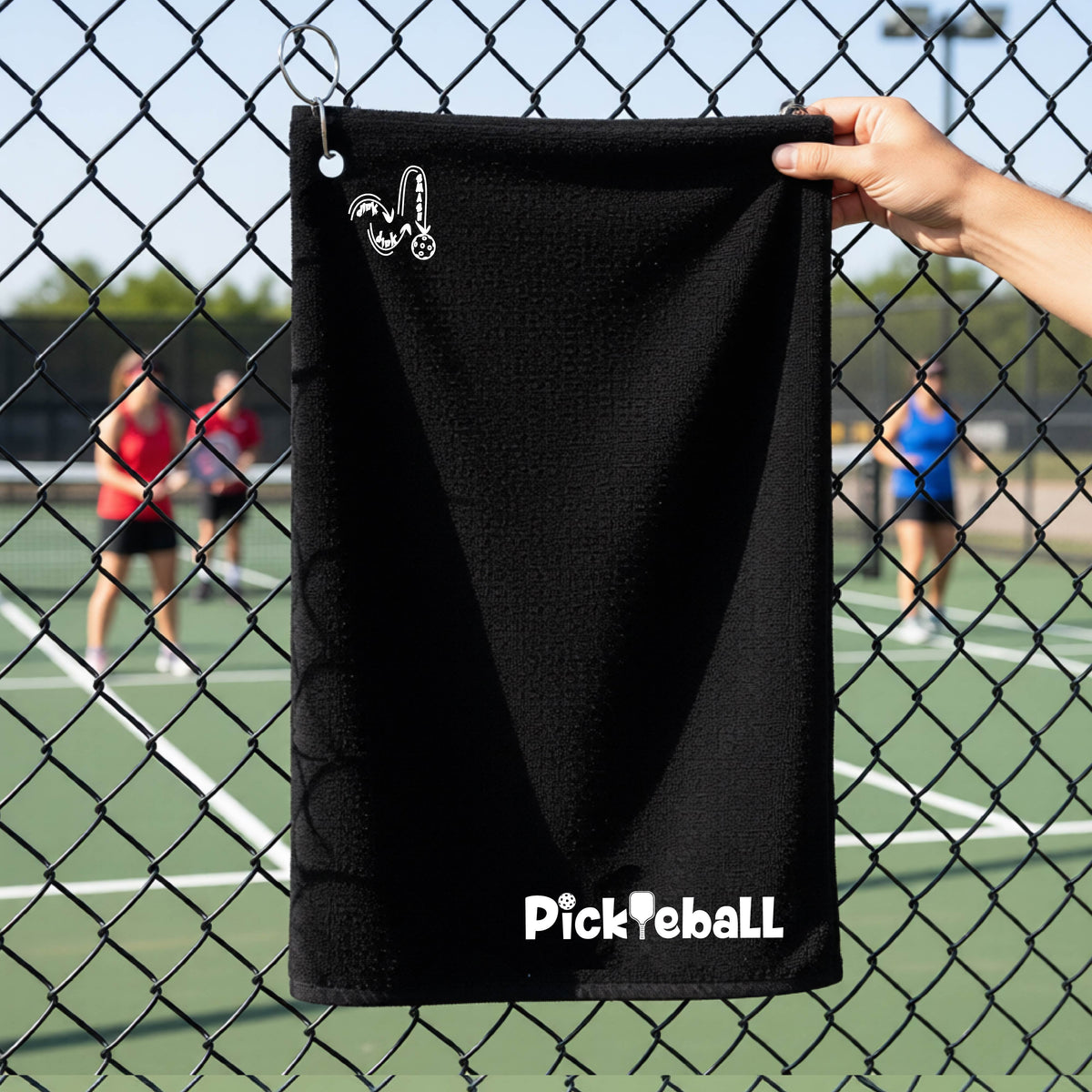 A close-up of a black terry-cloth court towel  with grommet and hook and a white graphic text 'PICKLEBALL' with a pickleball paddle replacing the L, on a pickleball court on the bottom right and a logo of ‘DINK DINK SMASH’ on the upper left of the towel.