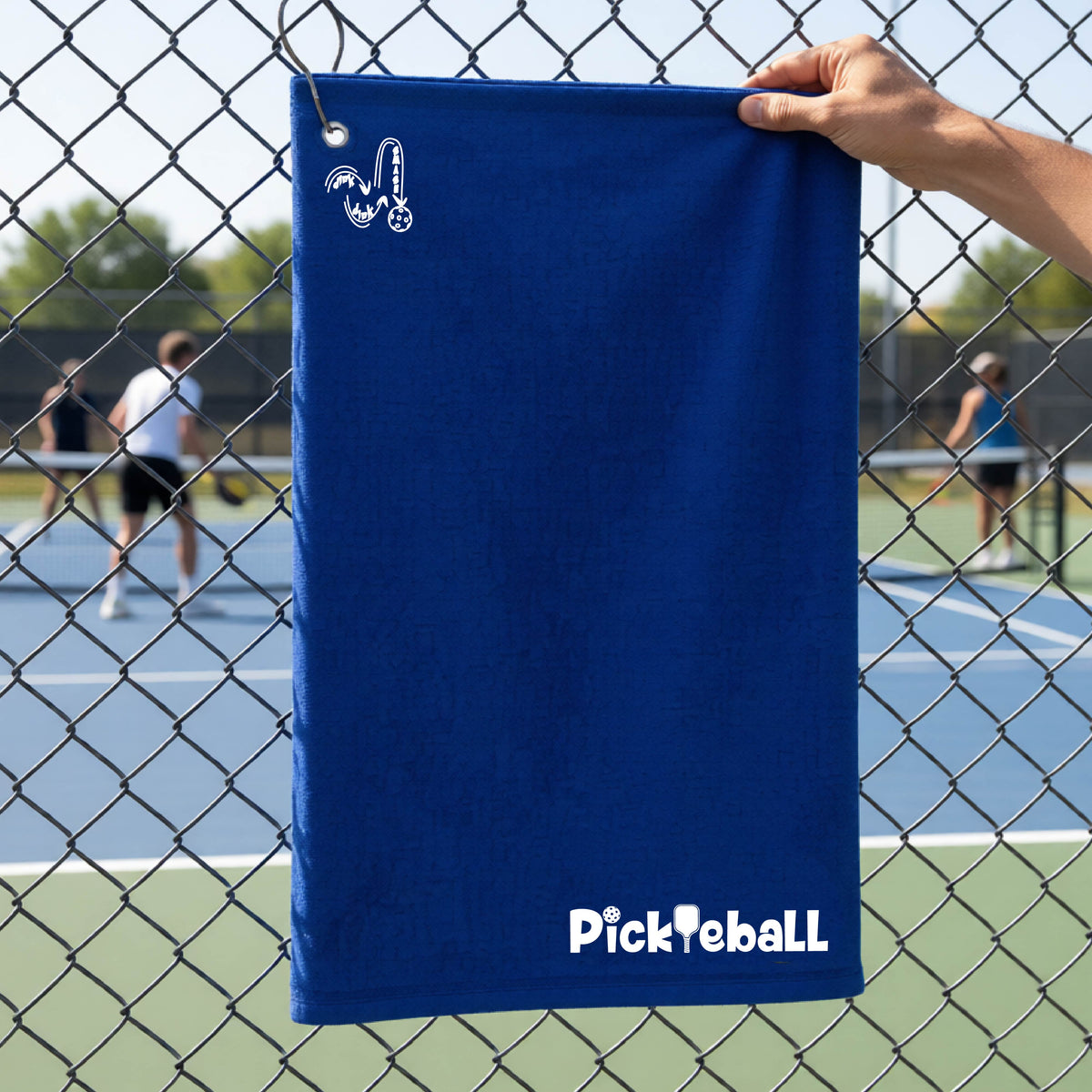 A close-up of a royal blue terry-cloth court towel  with grommet and hook and a white graphic text 'PICKLEBALL' with a pickleball paddle replacing the L, on a pickleball court on the bottom right and a logo of ‘DINK DINK SMASH’ on the upper left of the towel.