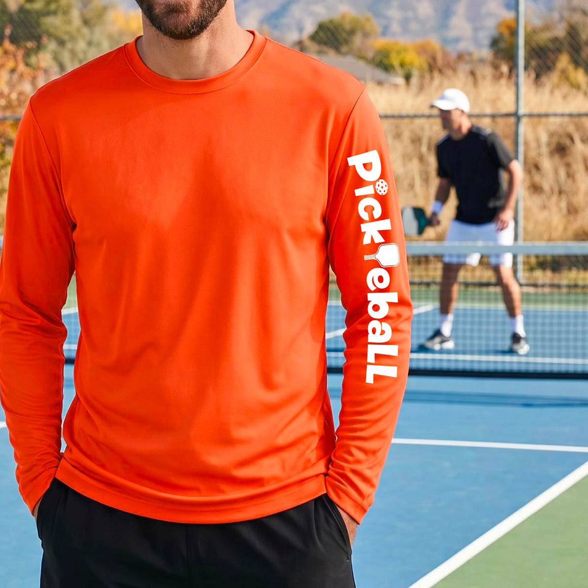 A man wearing a bright orange long-sleeve athletic shirt with 'Pickleball' printed vertically in white on the left sleeve, standing on a blue pickleball court with another player blurred in the background.