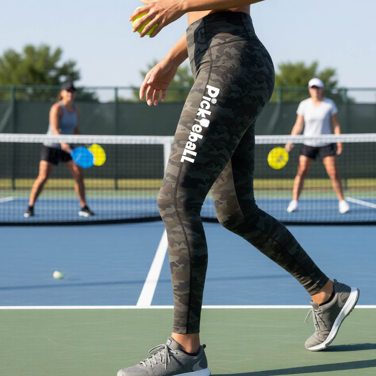 A close-up lifestyle shot of a woman on a pickleball court wearing high-waisted camo leggings with 'Pickleball' printed in white down the side of her leg.