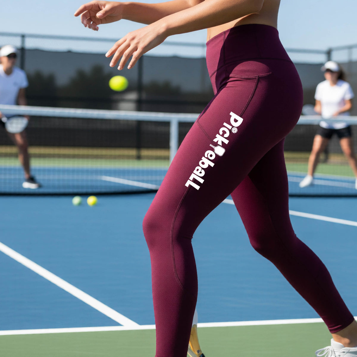 A close-up lifestyle shot of a woman on a pickleball court wearing high-waisted maroon leggings with 'Pickleball' printed in white down the side of her leg.