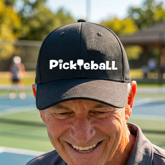 A close-up of a mid-profile black baseball cap with a white graphic text 'PICKLEBALL' with a pickleball paddle replacing the L, on a pickleball court.