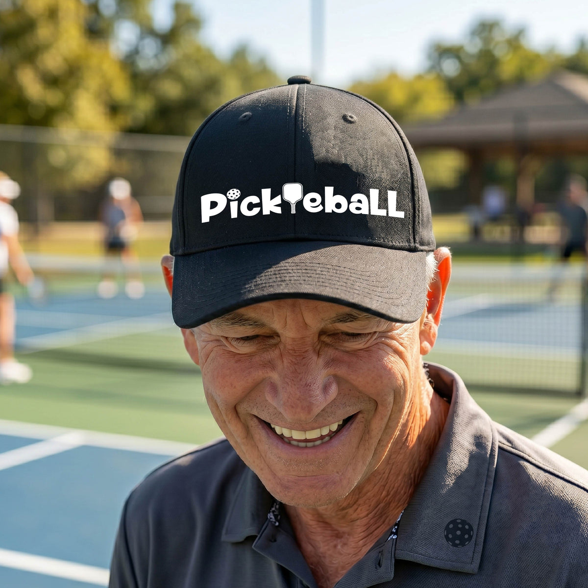 A close-up of a mid-profile black baseball cap with a white graphic text 'PICKLEBALL' with a pickleball paddle replacing the L, on a pickleball court.