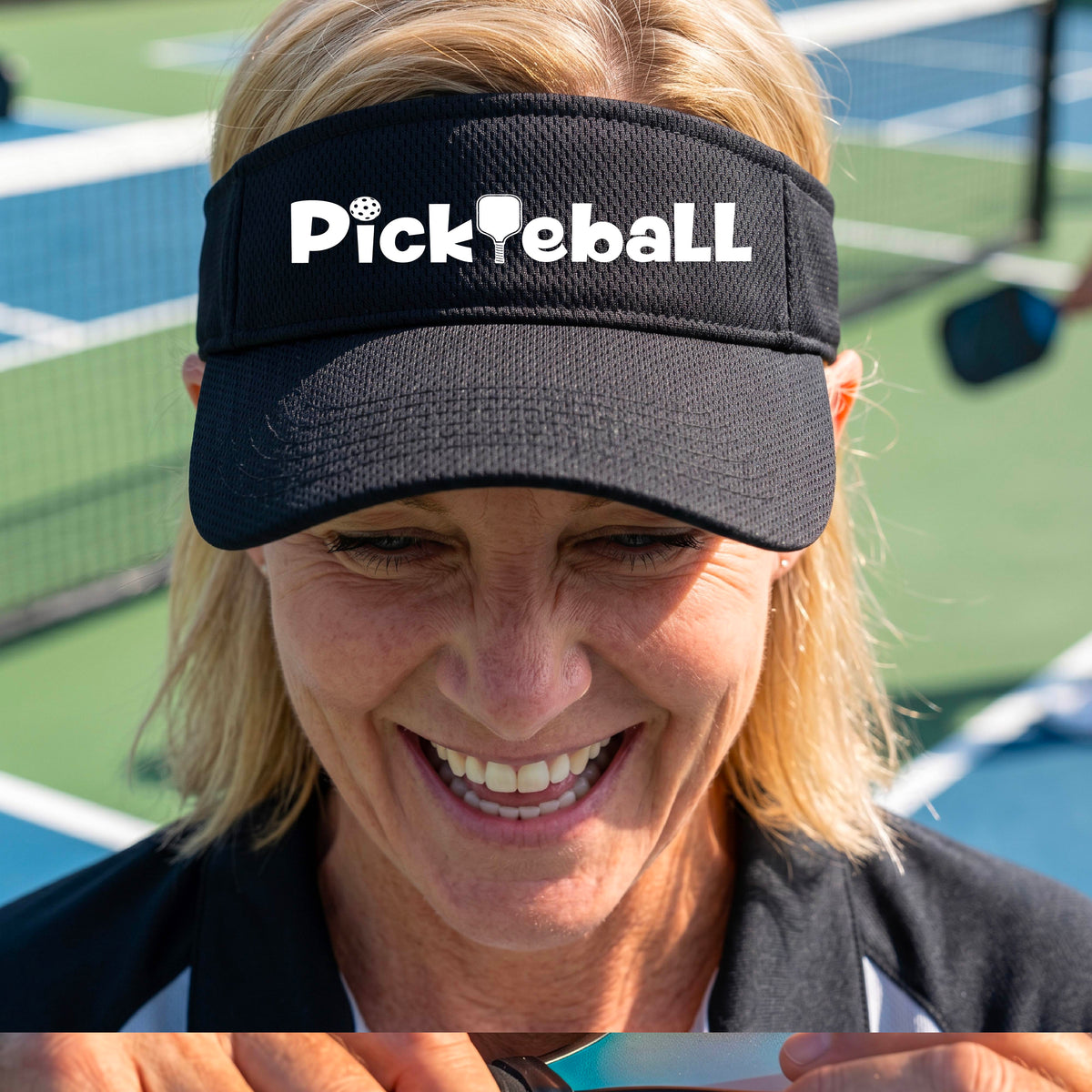 A close-up of a black athletic visor with a white graphic text 'PICKLEBALL' with a pickleball paddle replacing the L, on a pickleball court.