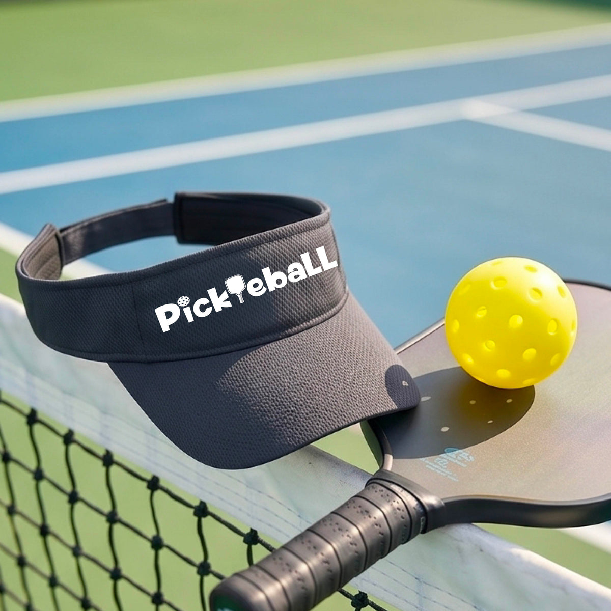 A close-up of a graphite gray athletic visor with a white graphic text 'PICKLEBALL' with a pickleball paddle replacing the L, on a pickleball court.