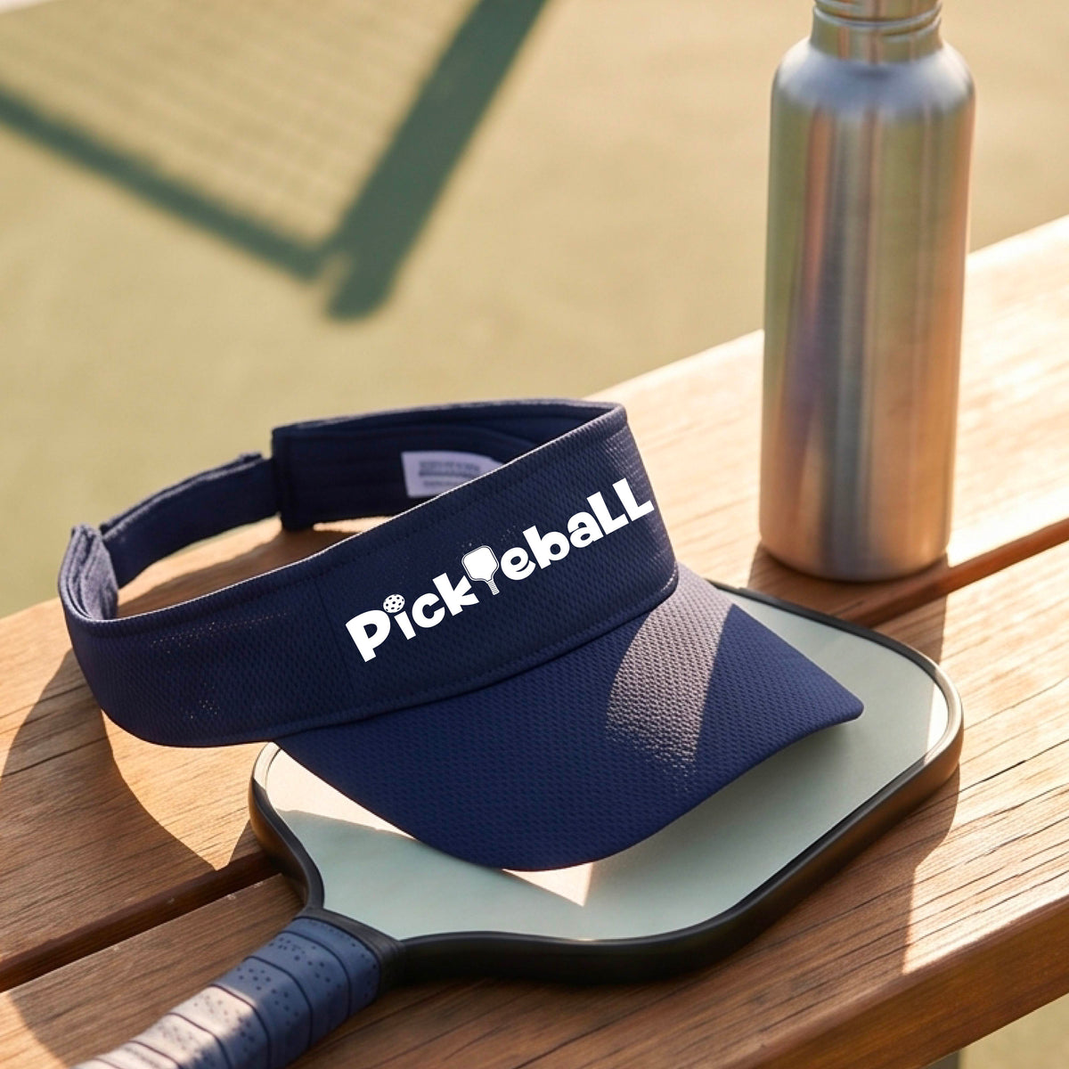 A close-up of a navy blue athletic visor with a white graphic text 'PICKLEBALL' with a pickleball paddle replacing the L, on a pickleball court.