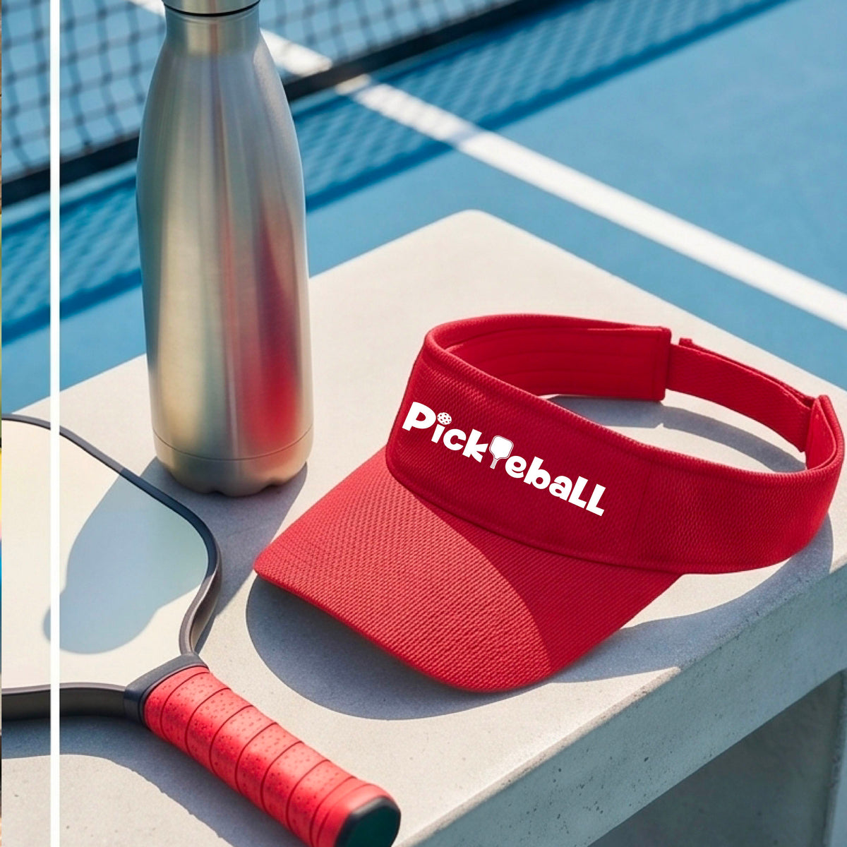 A close-up of a red athletic visor with a white graphic text 'PICKLEBALL' with a pickleball paddle replacing the L, on a pickleball court.