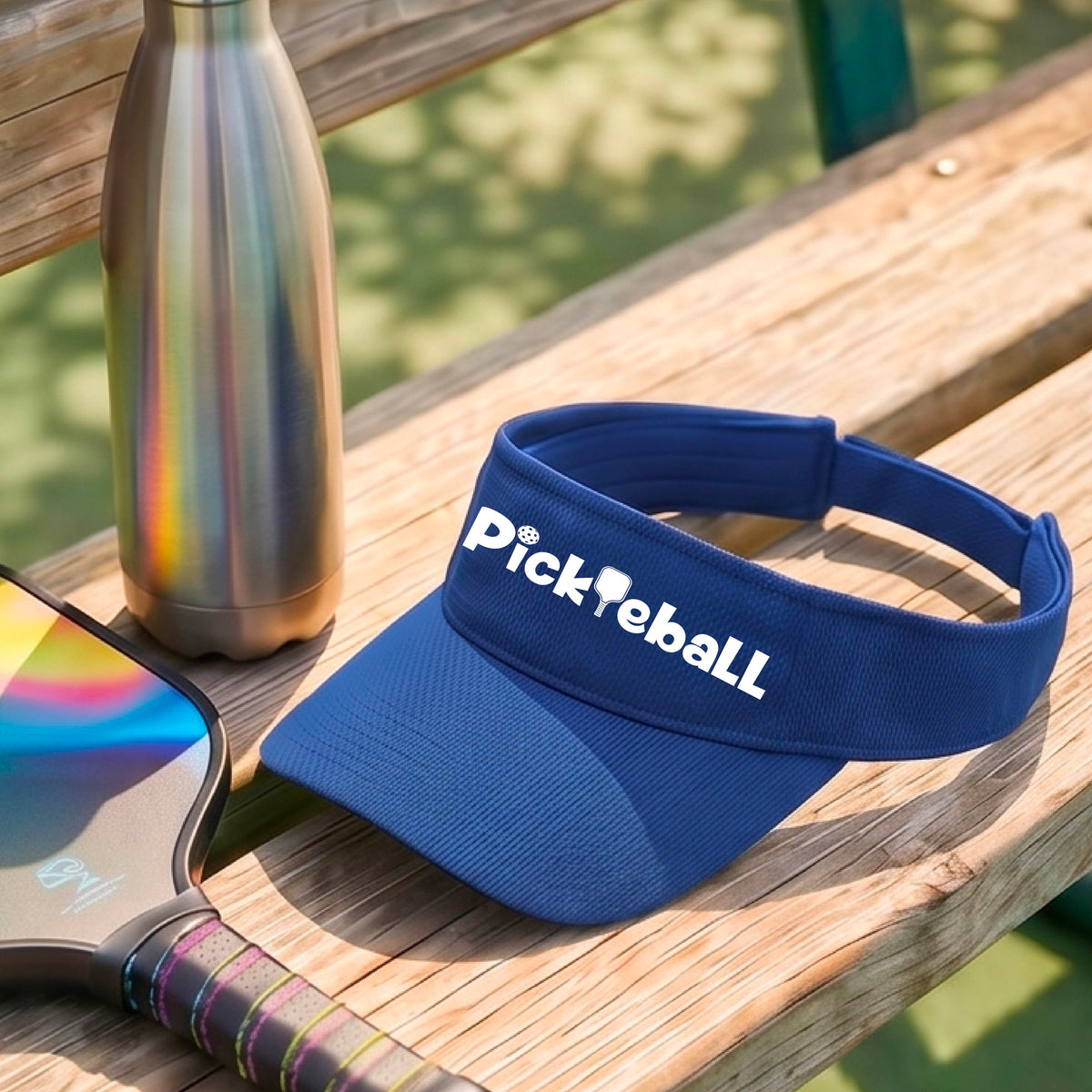 A close-up of a royal blue athletic visor with a white graphic text 'PICKLEBALL' with a pickleball paddle replacing the L, on a pickleball court.