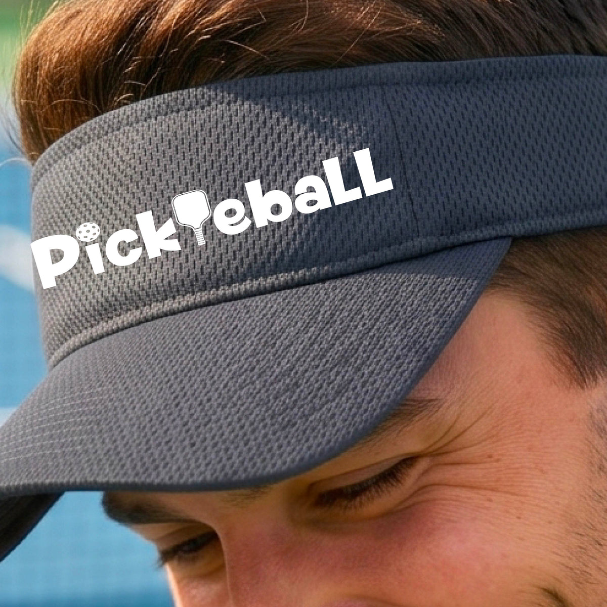 A close-up of a graphite gray athletic visor with a white graphic text 'PICKLEBALL' with a pickleball paddle replacing the L, on a pickleball court.