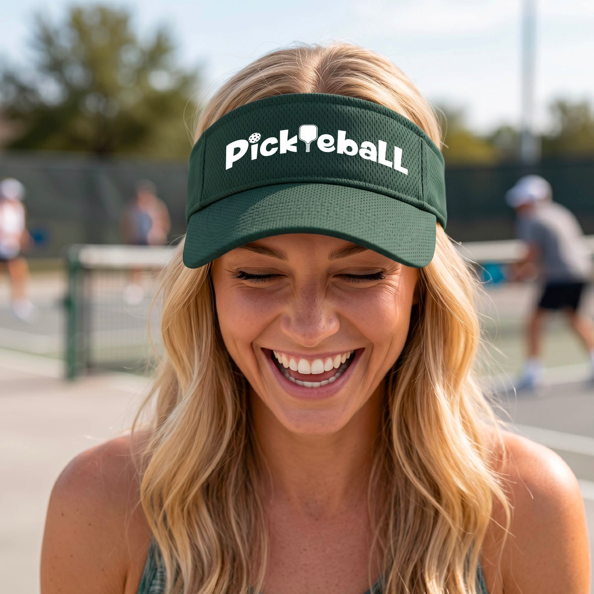 A close-up of a green athletic visor with a white graphic text 'PICKLEBALL' with a pickleball paddle replacing the L, on a pickleball court.