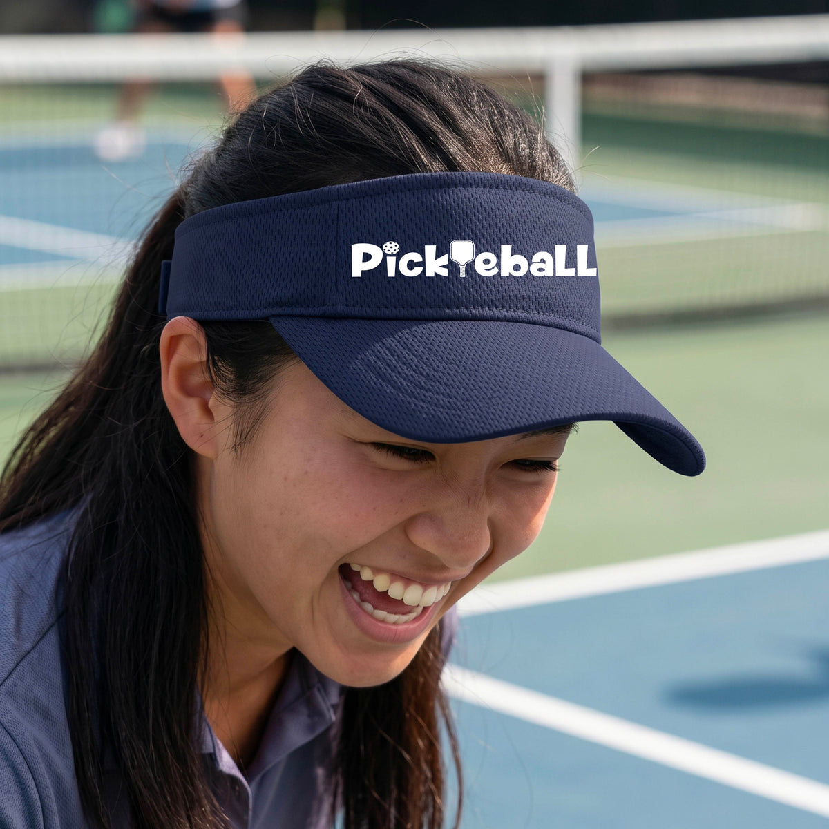 A close-up of a navy blue athletic visor with a white graphic text 'PICKLEBALL' with a pickleball paddle replacing the L, on a pickleball court.