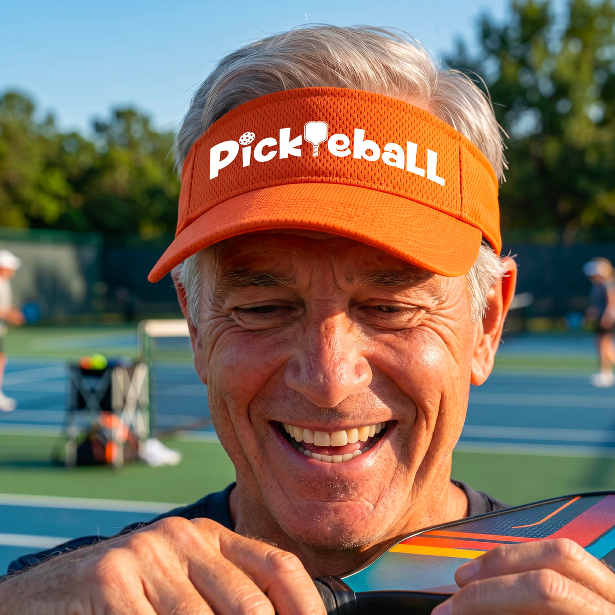 A close-up of an orange athletic visor with a white graphic text 'PICKLEBALL' with a pickleball paddle replacing the L, on a pickleball court.