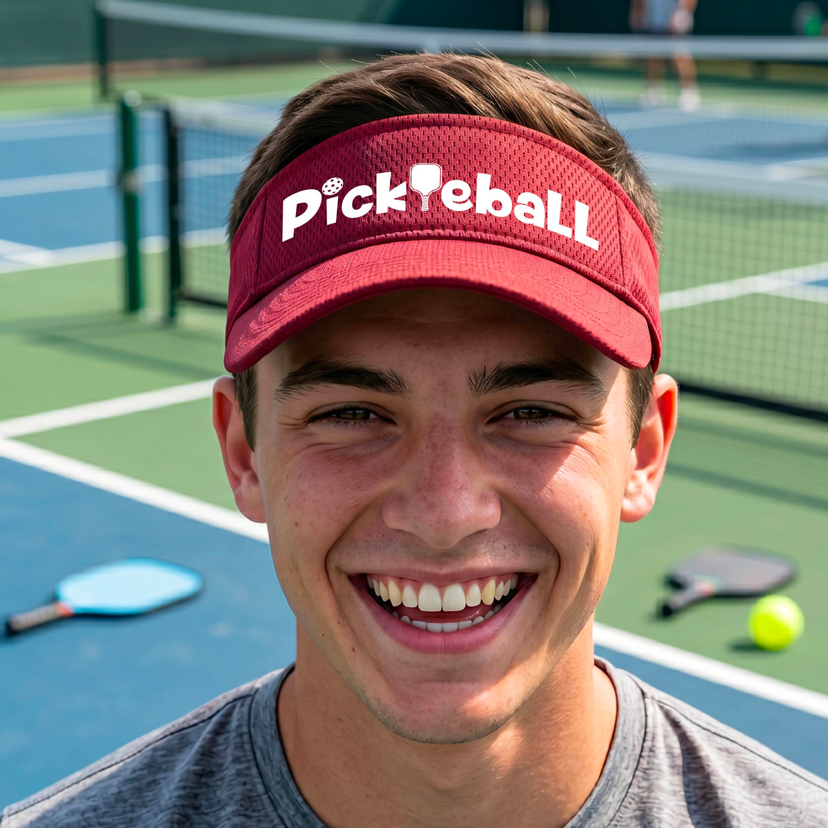 A close-up of a red athletic visor with a white graphic text 'PICKLEBALL' with a pickleball paddle replacing the L, on a pickleball court.