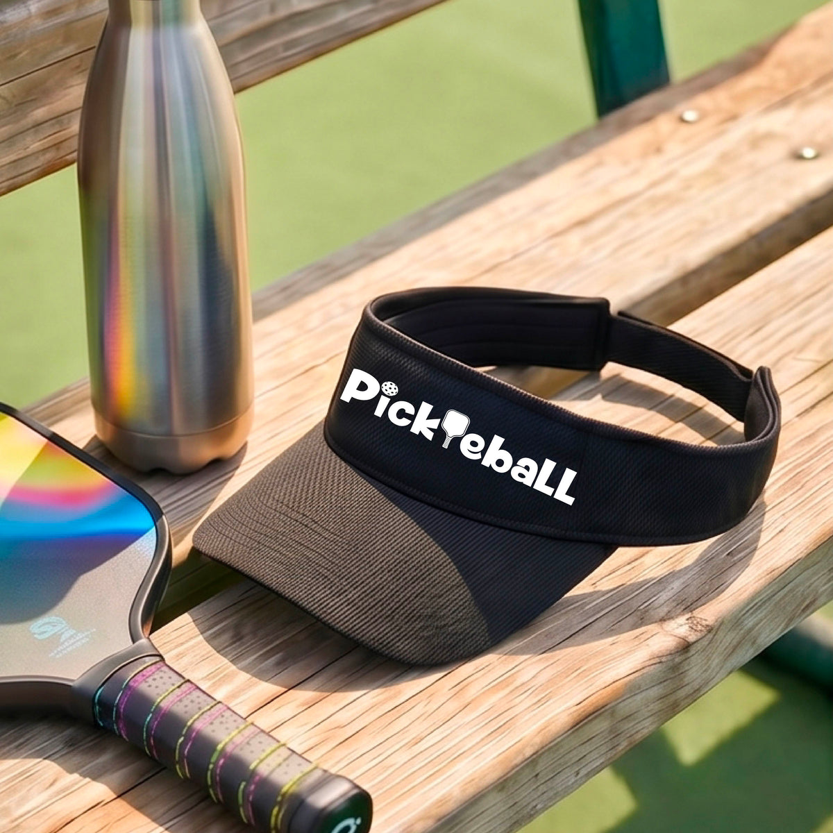 A close-up of a black athletic visor with a white graphic text 'PICKLEBALL'  with a pickleball paddle replacing the L, on a pickleball court.