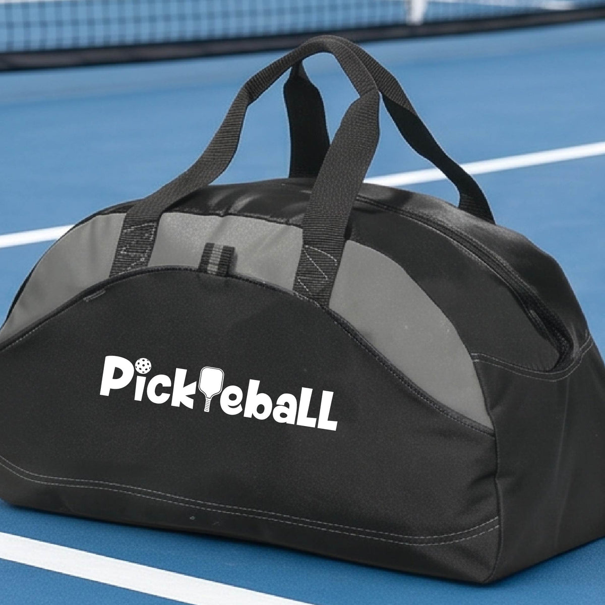 A close-up of a medium-size gray and black duffel bag with a white graphic text 'PICKLEBALL' on the leg with a pickleball paddle replacing the L, on a pickleball court.