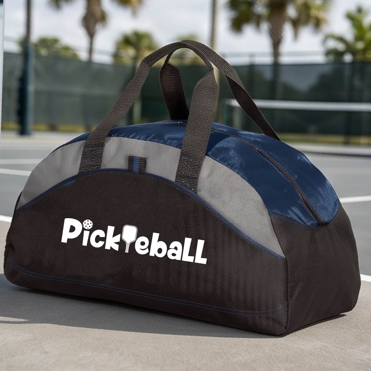 A close-up of a medium-size navy blue, gray and black duffel bag with a white graphic text 'PICKLEBALL' on the leg with a pickleball paddle replacing the L, on a pickleball court.