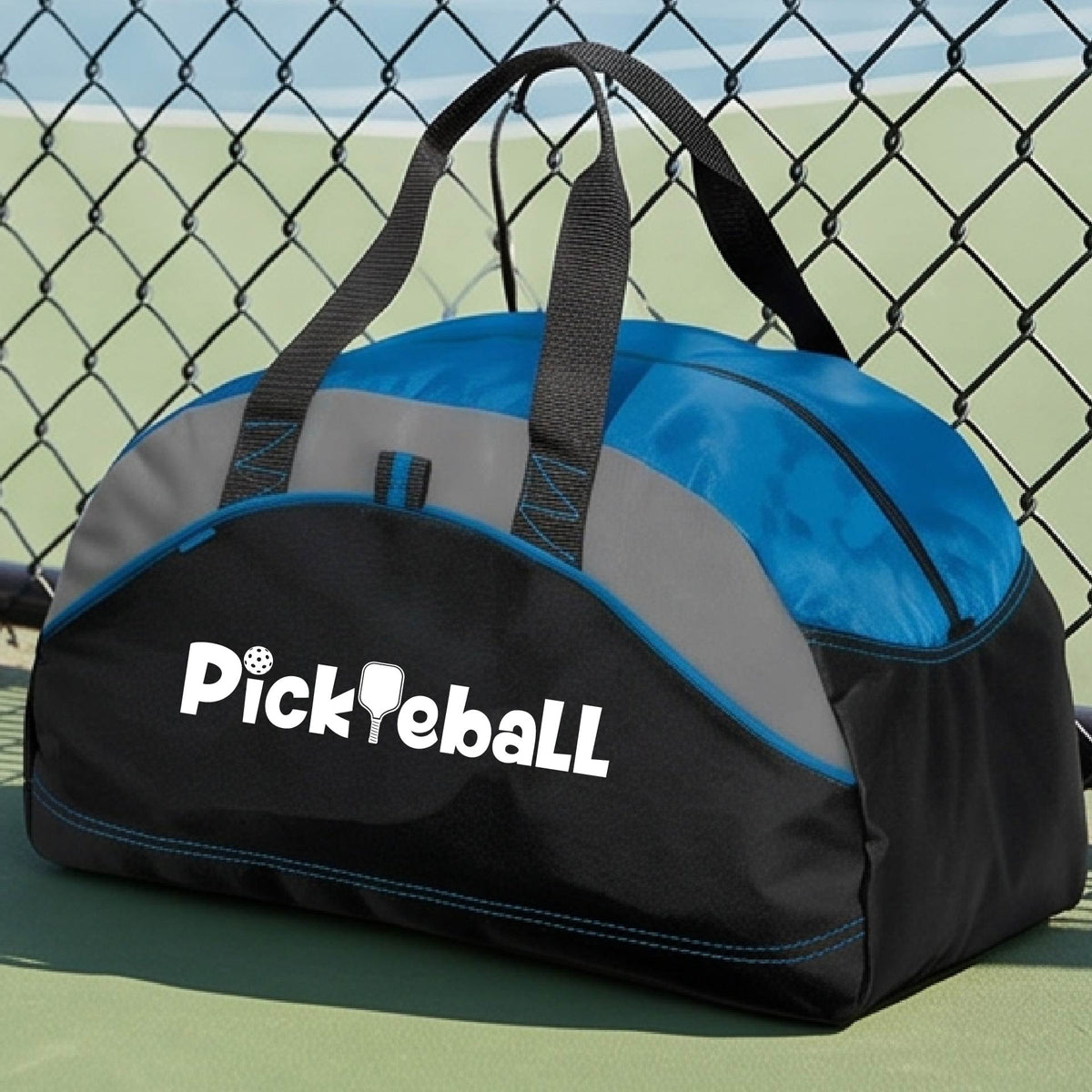 A close-up of a medium-size royal blue,  gray and black duffel bag with a white graphic text 'PICKLEBALL' on the leg with a pickleball paddle replacing the L, on a pickleball court.