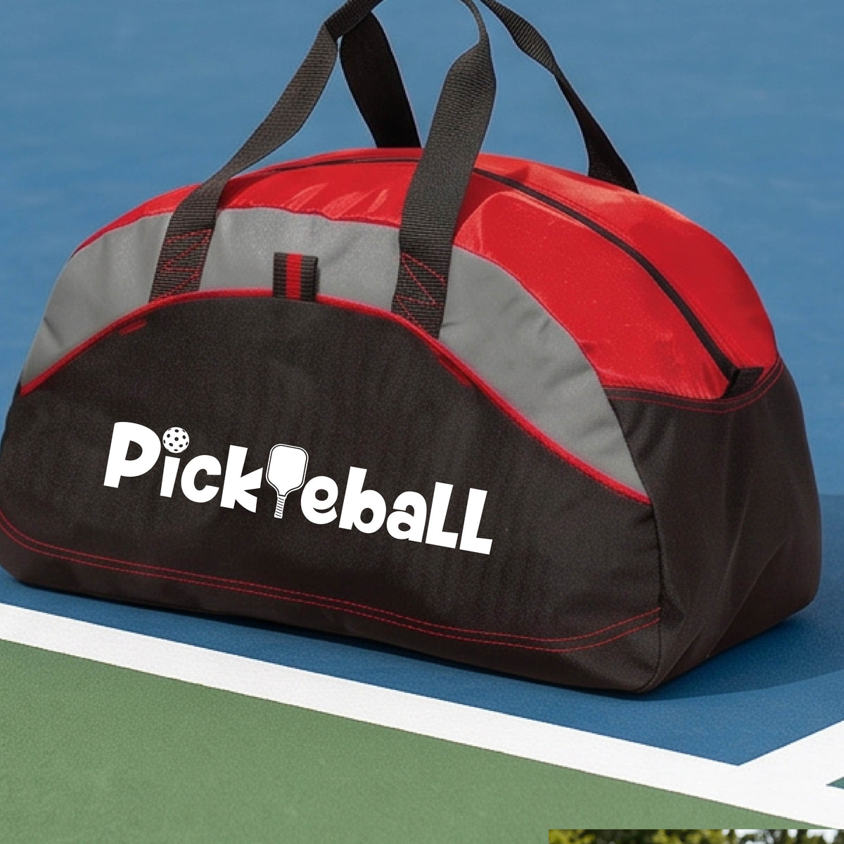 A close-up of a medium-size red, gray and black duffel bag with a white graphic text 'PICKLEBALL' on the leg with a pickleball paddle replacing the L, on a pickleball court.