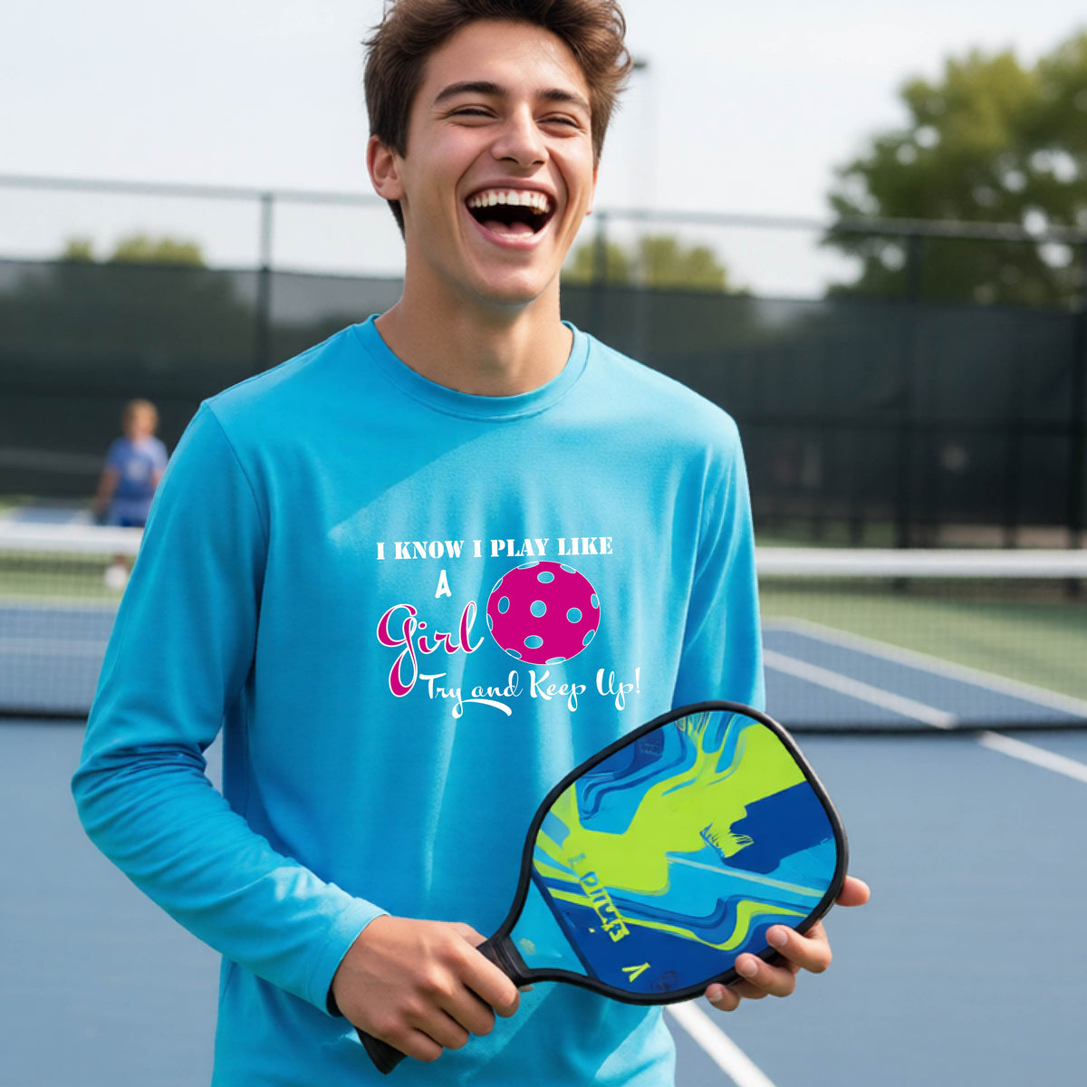 A young person laughing while wearing a bright blue long-sleeve t-shirt with a pink and white graphic that reads 'I KNOW I PLAY LIKE A Girl Try and Keep Up!' with a pink pickleball illustration.