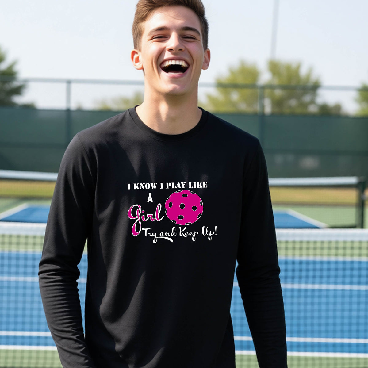 A young person laughing while wearing a black long-sleeve t-shirt with a pink and white graphic that reads 'I KNOW I PLAY LIKE A Girl Try and Keep Up!' with a pink pickleball illustration.