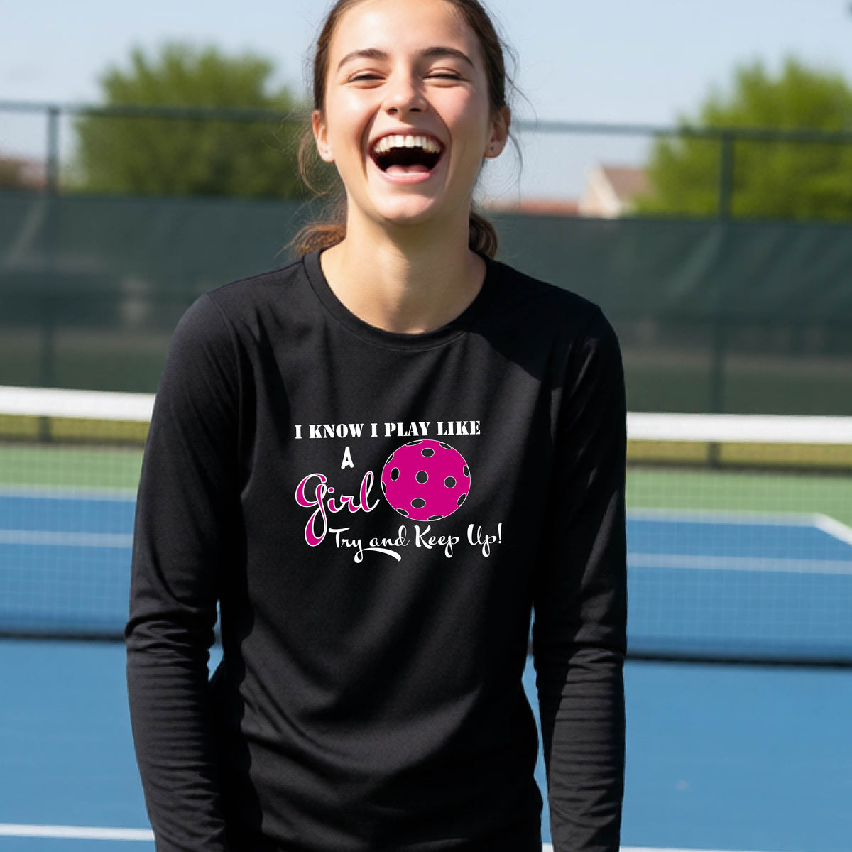 A young person laughing while wearing a black long-sleeve t-shirt with a pink and white graphic that reads 'I KNOW I PLAY LIKE A Girl Try and Keep Up!' with a pink pickleball illustration.