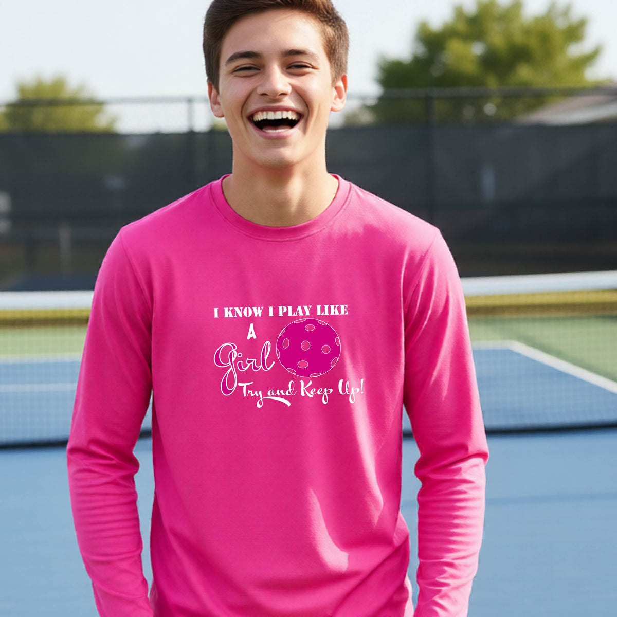 A young person laughing while wearing a pink long-sleeve t-shirt with a pink and white graphic that reads 'I KNOW I PLAY LIKE A Girl Try and Keep Up!' with a pink pickleball illustration.