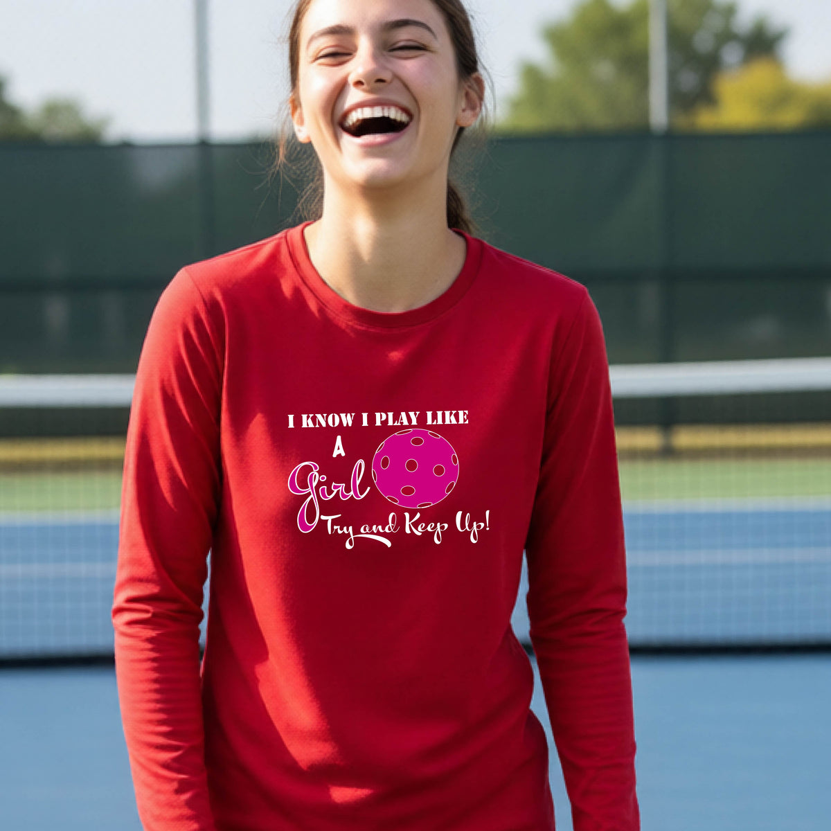 A young person laughing while wearing a red long-sleeve t-shirt with a pink and white graphic that reads 'I KNOW I PLAY LIKE A Girl Try and Keep Up!' with a pink pickleball illustration.
