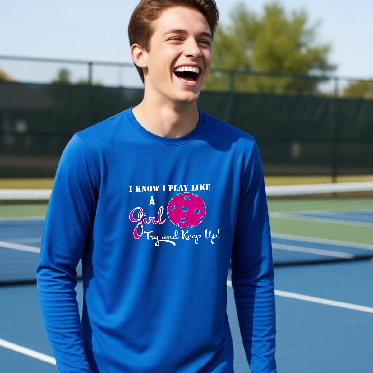 A young person laughing while wearing a royal blue long-sleeve t-shirt with a pink and white graphic that reads 'I KNOW I PLAY LIKE A Girl Try and Keep Up!' with a pink pickleball illustration.