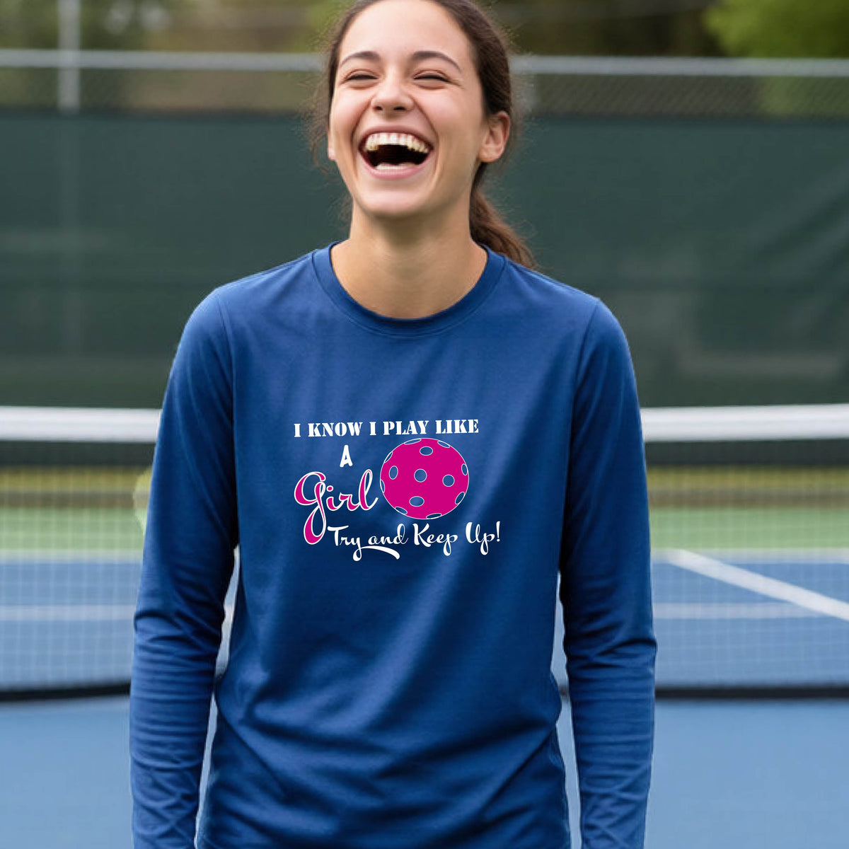 A young person laughing while wearing a royal blue long-sleeve t-shirt with a pink and white graphic that reads 'I KNOW I PLAY LIKE A Girl Try and Keep Up!' with a pink pickleball illustration.