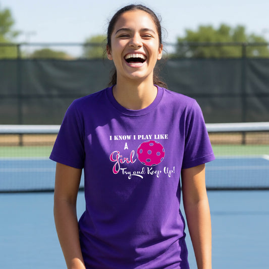 A close-up of a young person wearing a purple short-sleeve t-shirt  with a white graphic of ‘I KNOW I LAY LIKE A GIRL TRY AND KEEP UP’ in white and pink with a pink pickleball icon, in front of a pickleball court. 
